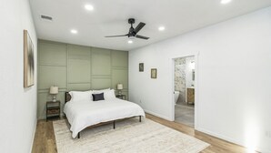 Ein Schlafzimmer mit Bett, Nachttischen, Deckenventilator und Blick ins Badezimmer.