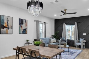 A modern living room with a dining area, featuring a chandelier, ceiling fan, abstract paintings, and a large window with curtains. - Stylish 4BR Home Family-friendly Near Downtown (San Antonio)