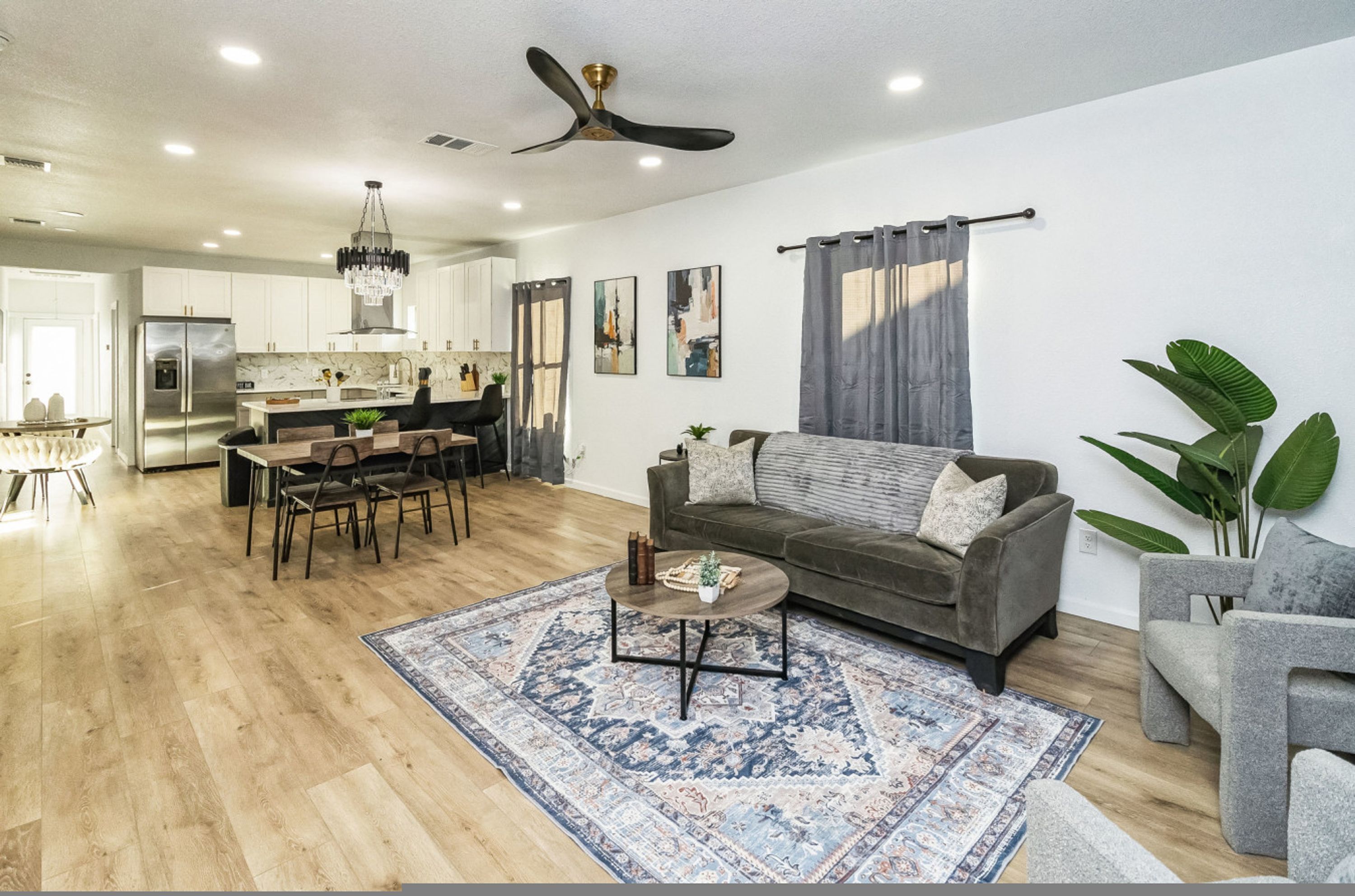 A modern living room with a sofa, coffee table, and a rug. There is a dining area with a table and chairs. The kitchen is visible in the background with cabinets and appliances.