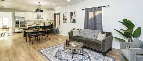 A modern living room with a sofa, coffee table, and a rug. There is a dining area with a table and chairs. The kitchen is visible in the background with cabinets and appliances.