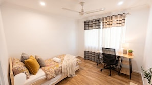 A small, well-lit room with a bed, a desk, and a chair.