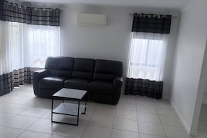 A living room with a black leather sofa, a small table, and two windows with black curtains.
