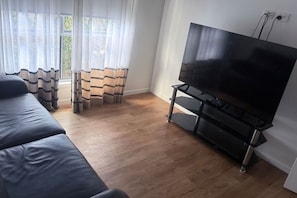 Olohuoneessa on musta sohva, mustalla jalustalla oleva televisio, puulattia ja ikkuna, jossa on valkoiset verhot.