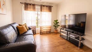 Un salon avec un canapé en cuir, un téléviseur à écran plat posé sur un meuble noir, une plante en pot et une fenêtre avec des rideaux.