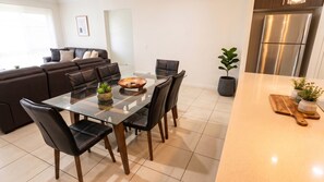A modern kitchen with a glass dining table, black chairs, a stainless steel refrigerator, and a potted plant.