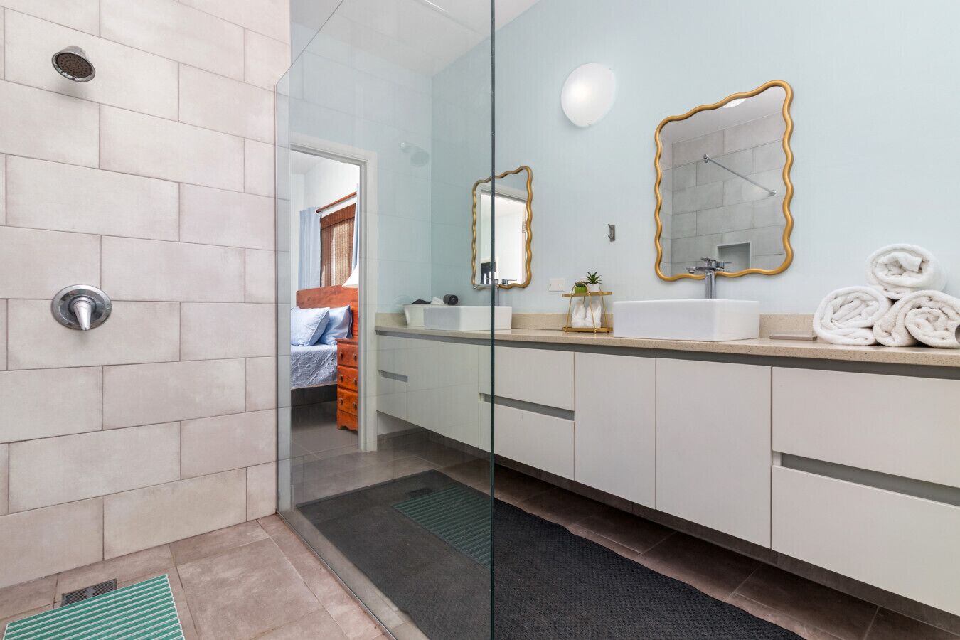 A modern bathroom with a glass shower enclosure, a white sink with gold-framed mirrors, and a visible bedroom with a bed and a door leading to another room.