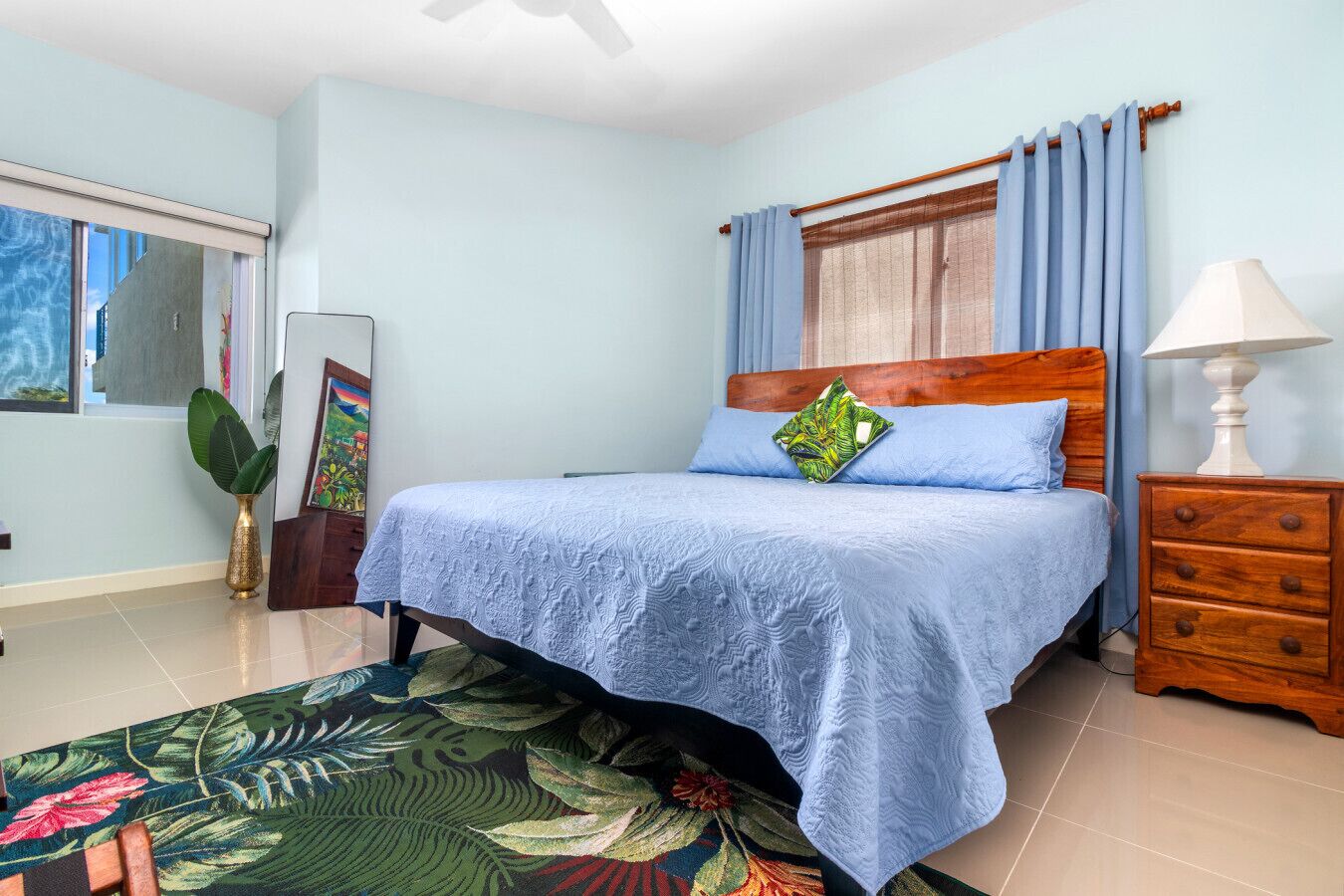 A bedroom with a bed, a nightstand, a lamp, a window with curtains, a painting, and a plant.