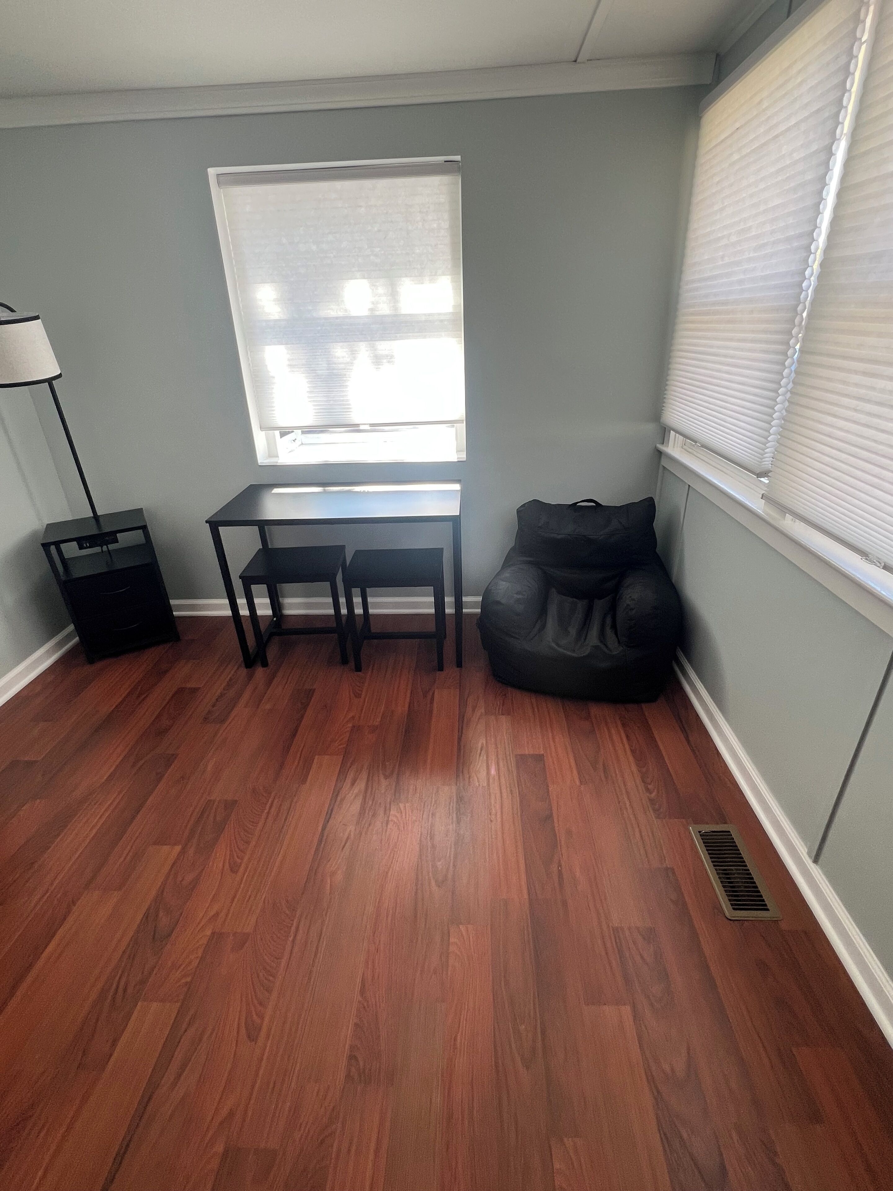 A room with a desk, two chairs, a black bean bag, a floor lamp, and a window with blinds.