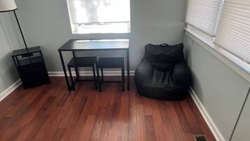 A room with a desk, two chairs, a black bean bag, a floor lamp, and a window with blinds.
