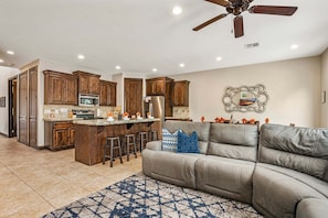 A modern kitchen with wooden cabinets, a central island, and a dining area with a gray sofa and patterned rug.