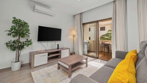 A modern living room with a flat-screen TV, a wooden coffee table, a grey sofa with a yellow pillow, a potted plant, and a sliding glass door leading to a balcony.