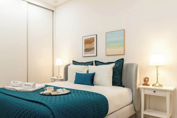 Une chambre d’hôtel comprenant un lit, deux tables de chevet, une petite table, un plateau avec une tasse et une assiette, et deux lampes fixées au mur.