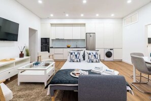 Une pièce moderne et bien éclairée, comprenant un lit, une table basse, un téléviseur, un coin repas et une cuisine.