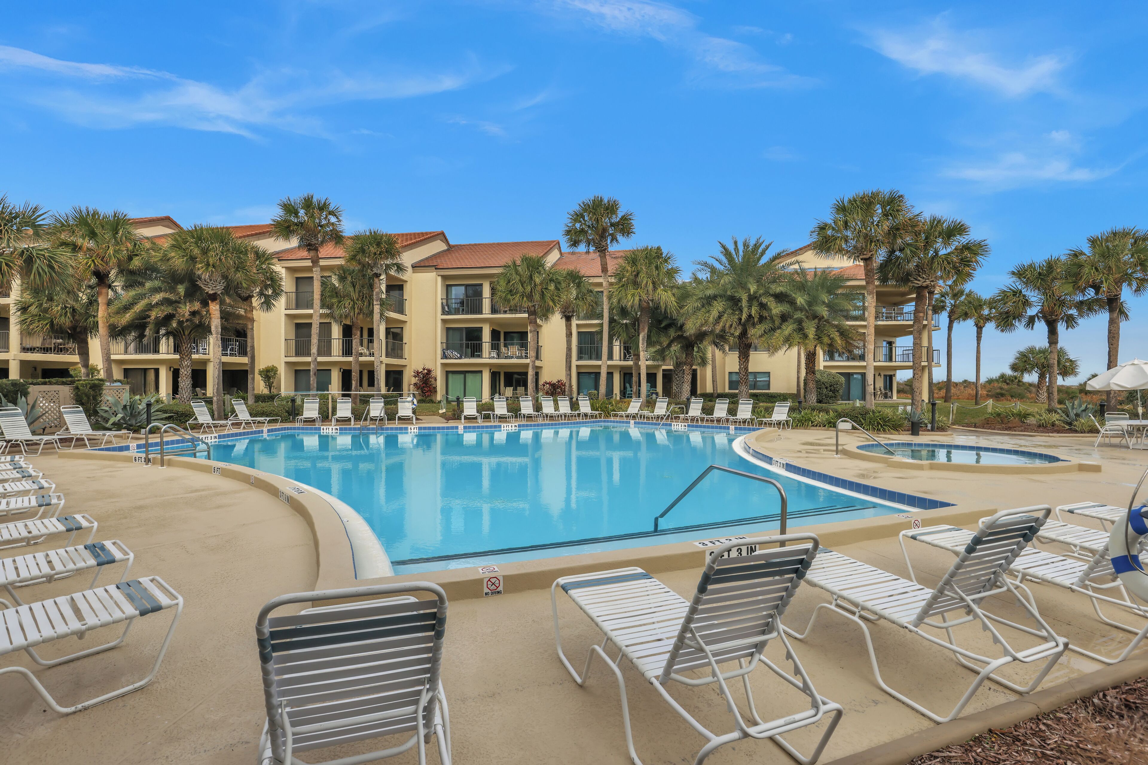 A pool area with lounge chairs and palm trees, surrounded by a multi-story building with balconies.