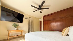 Un dormitorio con cama, cabecera de madera, ventilador de techo, televisor montado en la pared y una mesita con una planta.