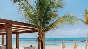 Una terraza de madera frente a la playa con sillas y una mesa blancas, palmeras y una vista despejada del océano.