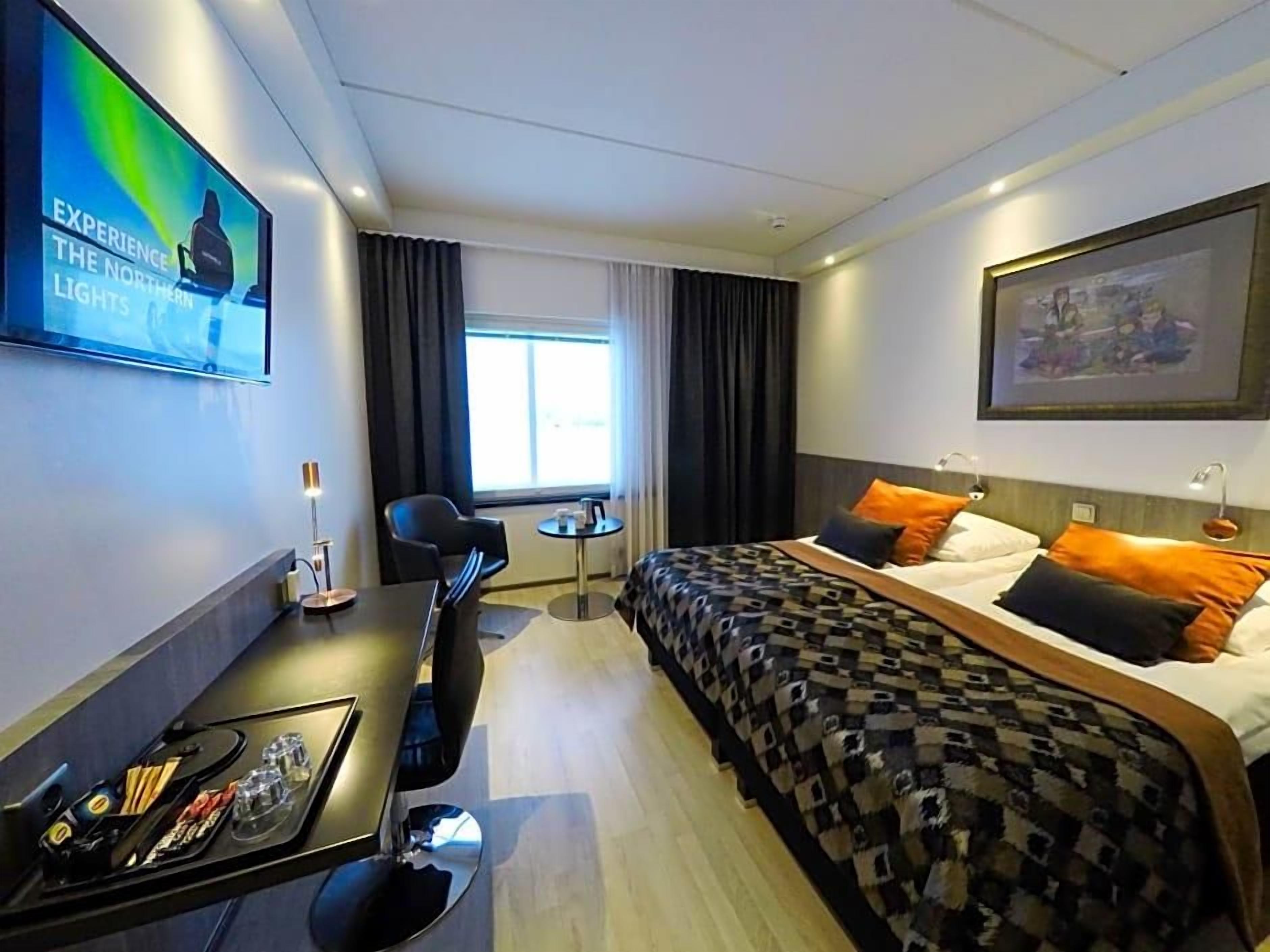 A hotel room with a bed, a desk with a chair, a small table, a TV, and a window with curtains.
