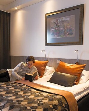 A person reading a book on a bed with a patterned blanket, in a room with a framed picture on the wall and two bedside lamps. - Inari Hotel (Inari)