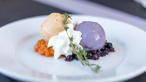 Une assiette à dessert avec une boule de glace, une sphère violette et une quenelle de crème, le tout garni de baies et d’herbes aromatiques.
