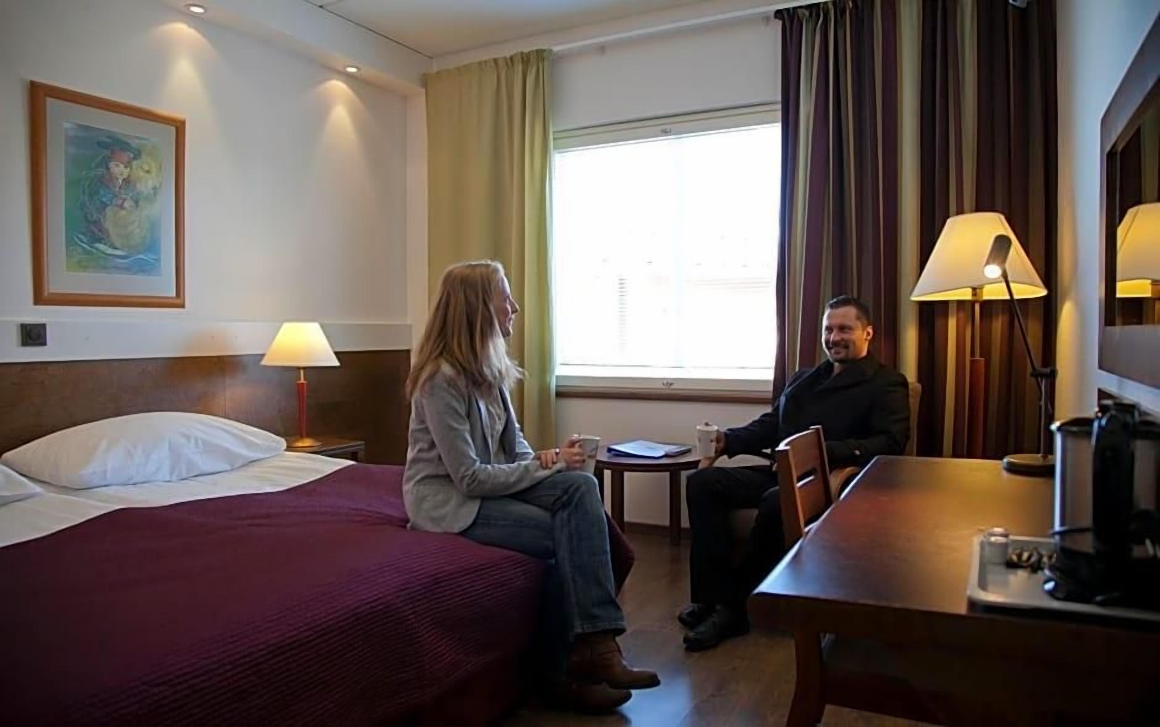 A hotel room with a bed, a small table, a chair, a lamp, and a window with curtains.