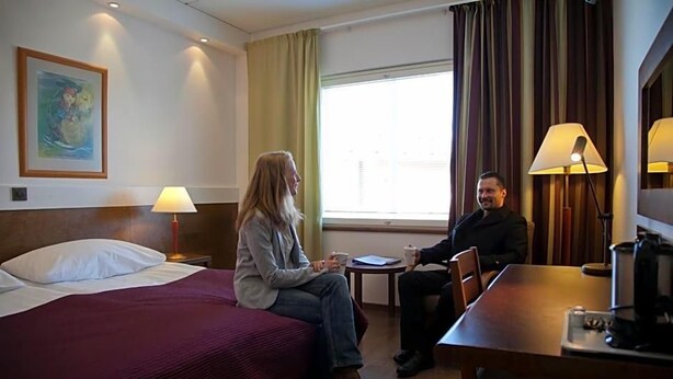Une chambre d’hôtel avec un lit, une petite table, une chaise, une lampe et une fenêtre avec des rideaux.