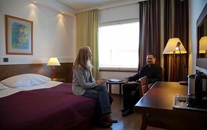 A hotel room with a bed, a small table, a chair, a lamp, and a window with curtains. - Inari Hotel (Inari)