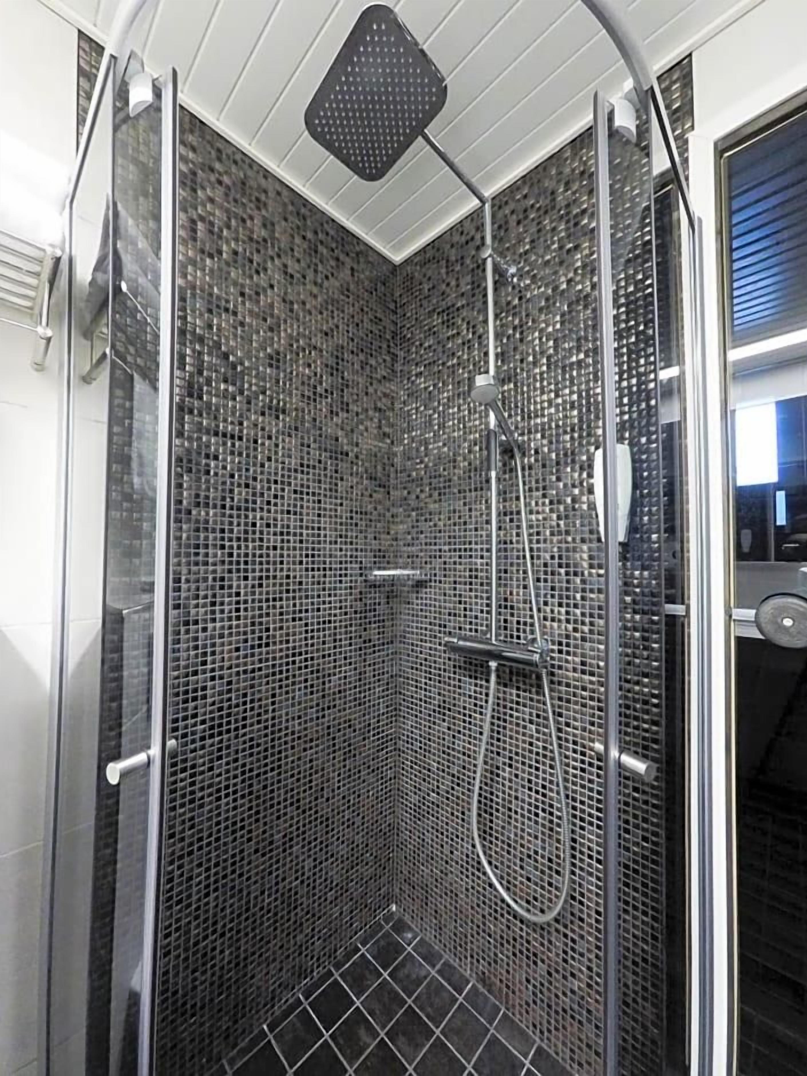 Una ducha con paredes de vidrio, un muro con mosaicos de azulejos, un cabezal de ducha manual y una boquilla de ducha manual.