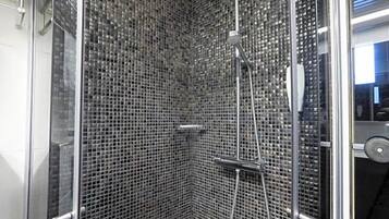 Una ducha con paredes de vidrio, un muro con mosaicos de azulejos, un cabezal de ducha manual y una boquilla de ducha manual.