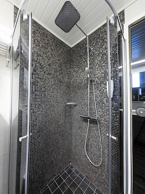 Una ducha con paredes de vidrio, un muro con mosaicos de azulejos, un cabezal de ducha manual y una boquilla de ducha manual.