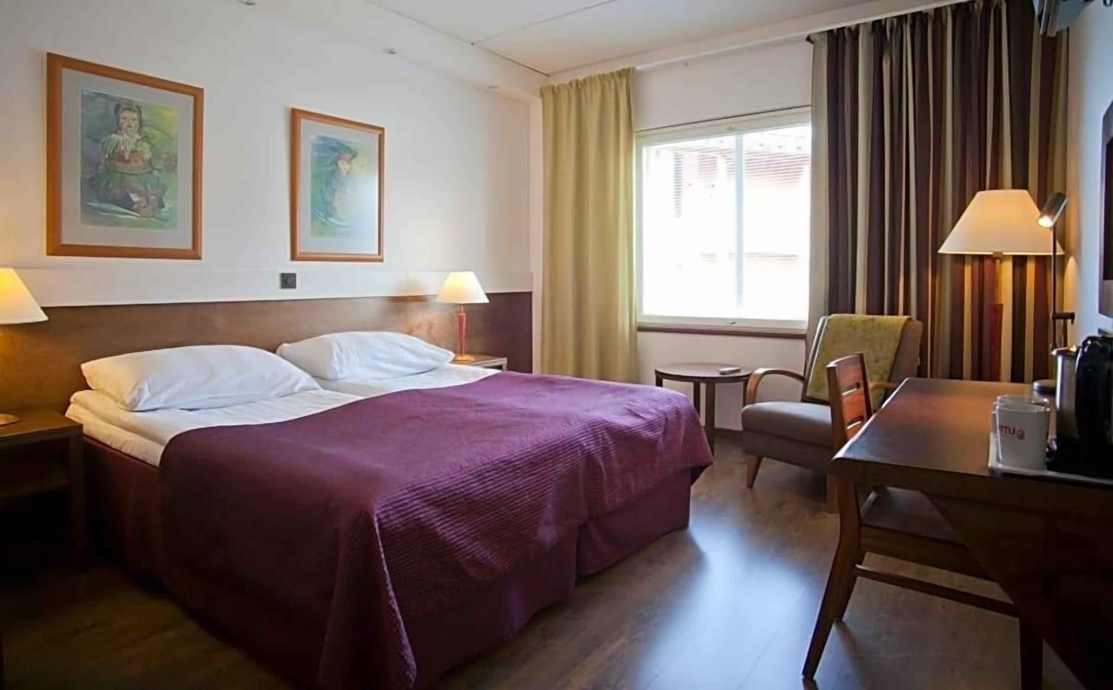 A hotel room with a bed, two paintings on the wall, a chair, a table, and a window with curtains.