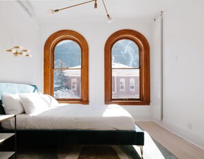 Une chambre à coucher avec un lit, deux grandes fenêtres en arc de cercle, un luminaire au plafond et une vue sur un bâtiment enneigé.