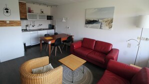 Un espace de vie compact comprenant un coin cuisine, un coin repas, un canapé rouge, un fauteuil en osier et une petite table.