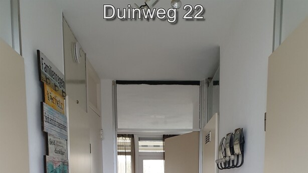 Un passadís amb una porta que dona a una habitació amb una cadira vermella, un llum fixada a la paret i una sèrie de quadres emmarcats a la pared esquerra.