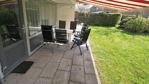 A patio with a table and chairs, a glass door, and a grassy area with a hedge. - Comfort Vikingpark 25 (Westkapelle)