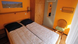 A room with two beds, a chair, a shelf with a lamp, and a painting on the wall.