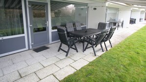 Een buitenterras met een tafel en stoelen, een overdekt gedeelte en een glazen deur naar een overdekte ruimte.