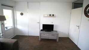 Een kamer met een deur, een televisie op een houten kast, een klok aan de muur en een raam met zonwering.