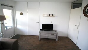 Ein Zimmer mit Tür, Fenster, einem Fernseher auf einem Sockel, einem Bücherregal und einer Uhr an der Wand.