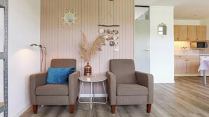 Une pièce comprenant deux fauteuils, une petite table ronde, un vase avec des plantes séchées, un miroir fixé au mur et, au fond, un coin cuisine.