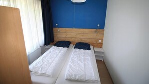 A small, single-room hotel room with a wooden headboard, two beds with white bedding, a window with white curtains, and a blue wall.