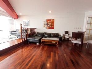 A living room with a wooden floor, a black leather sofa, a glass coffee table, and a wooden chair.