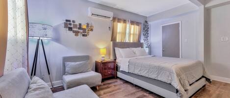 A bedroom with a bed, a chair, a bedside table, an air conditioning unit, and a wall with decorative items.
