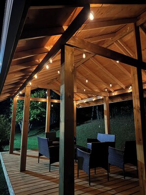 Een terras van hout met stoelen en tafels, onder een houten pergola met lichtslingers.