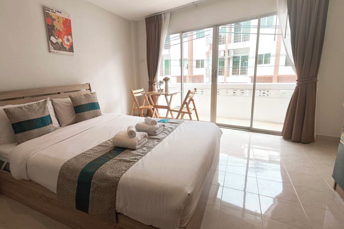 A hotel room with a bed, a small table with chairs, a window with curtains, and a poster on the wall.