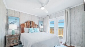Une chambre avec un grand lit, un ventilateur de plafond, une tête de lit en bois, une table de chevet avec une lampe et une vue sur l’extérieur grâce à une porte coulissante.