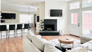 A modern living room with a fireplace, a ceiling fan, and a kitchen area in the background. - Big Backyard w/pool, Centrally Located (The Colony)