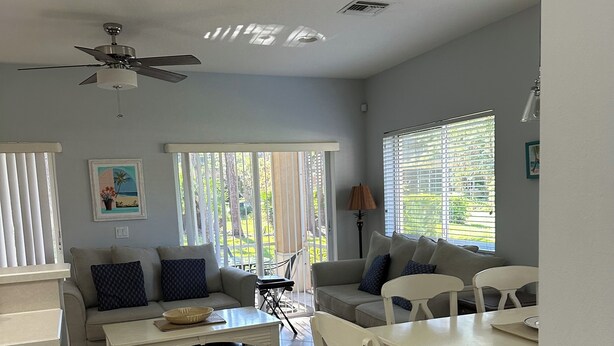 A spacious living area with a sofa, dining table, and chairs. There is a ceiling fan, a window with blinds, and a trash bin.