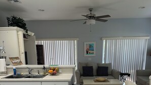 A modern kitchen with white cabinets, a sink, and a fruit bowl. There is a dining area with a table and chairs, and a ceiling fan.