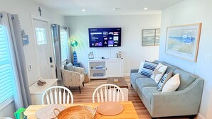 A modern living room with a dining area, a grey sofa, and a flat-screen TV mounted on the wall. - The Breakaway Beach Condo (North Wildwood)