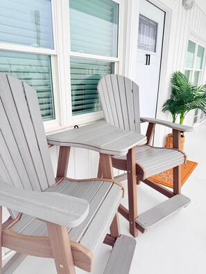 A set of Adirondack chairs with a wooden frame and grey cushions, positioned on a porch with a white door and windows in the background.